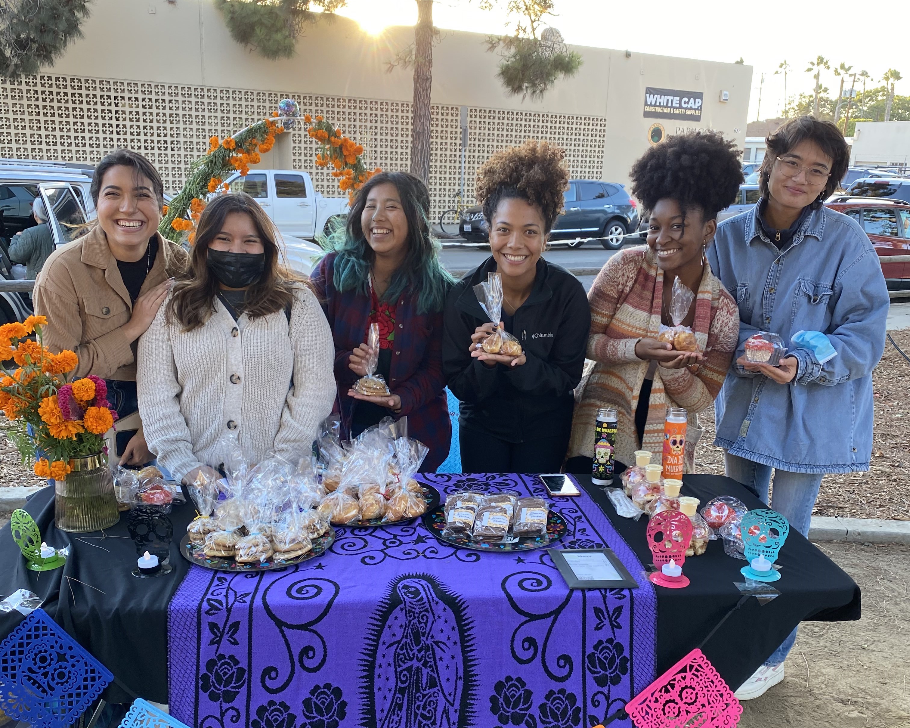 "Six students standing outside holding baked goods in front of table celebrating Dios De Los Muertos"
