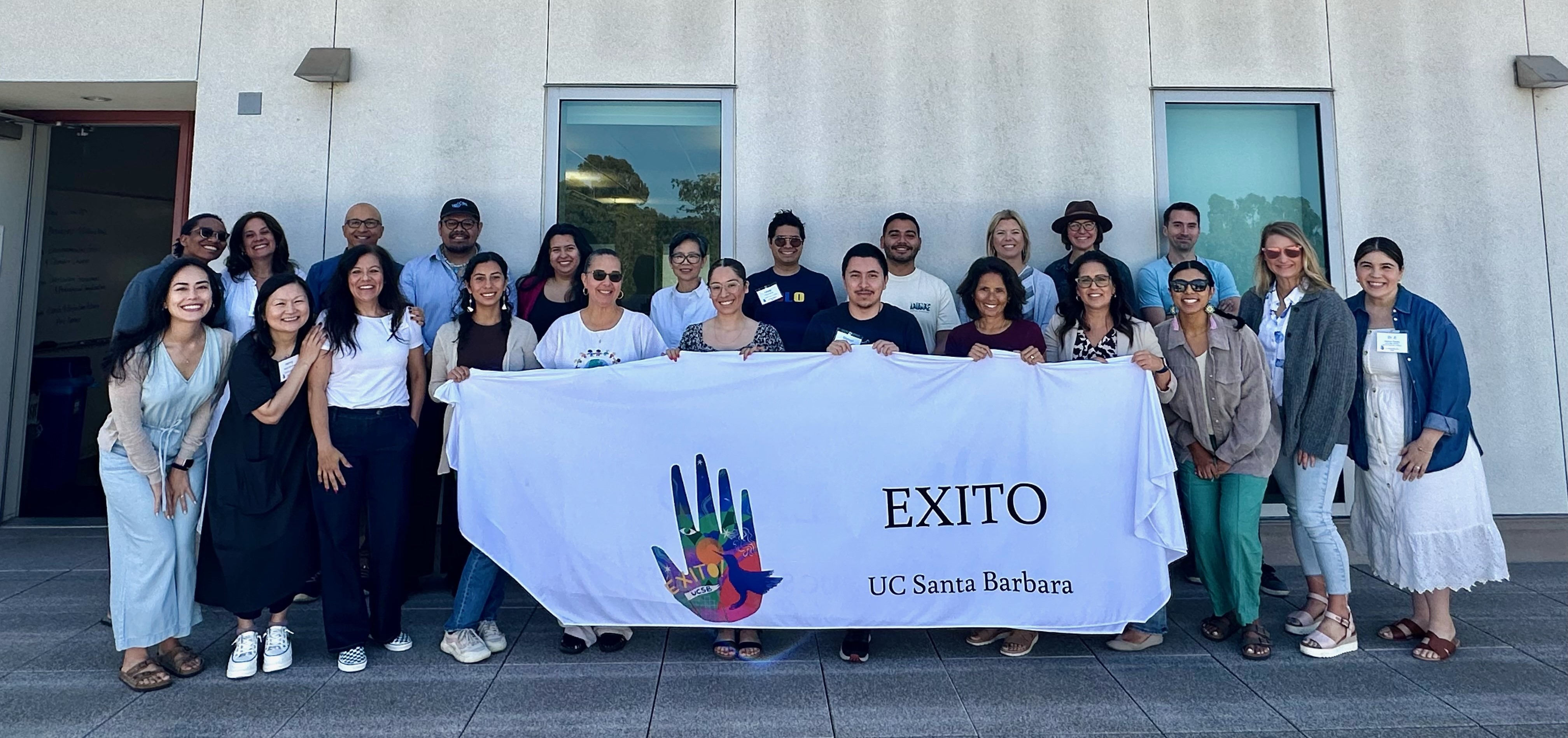 "A group of 23 teachers who participated in the 2024 EXITO Summer Institute smile while holding a white banner featuring the EXITO logo""