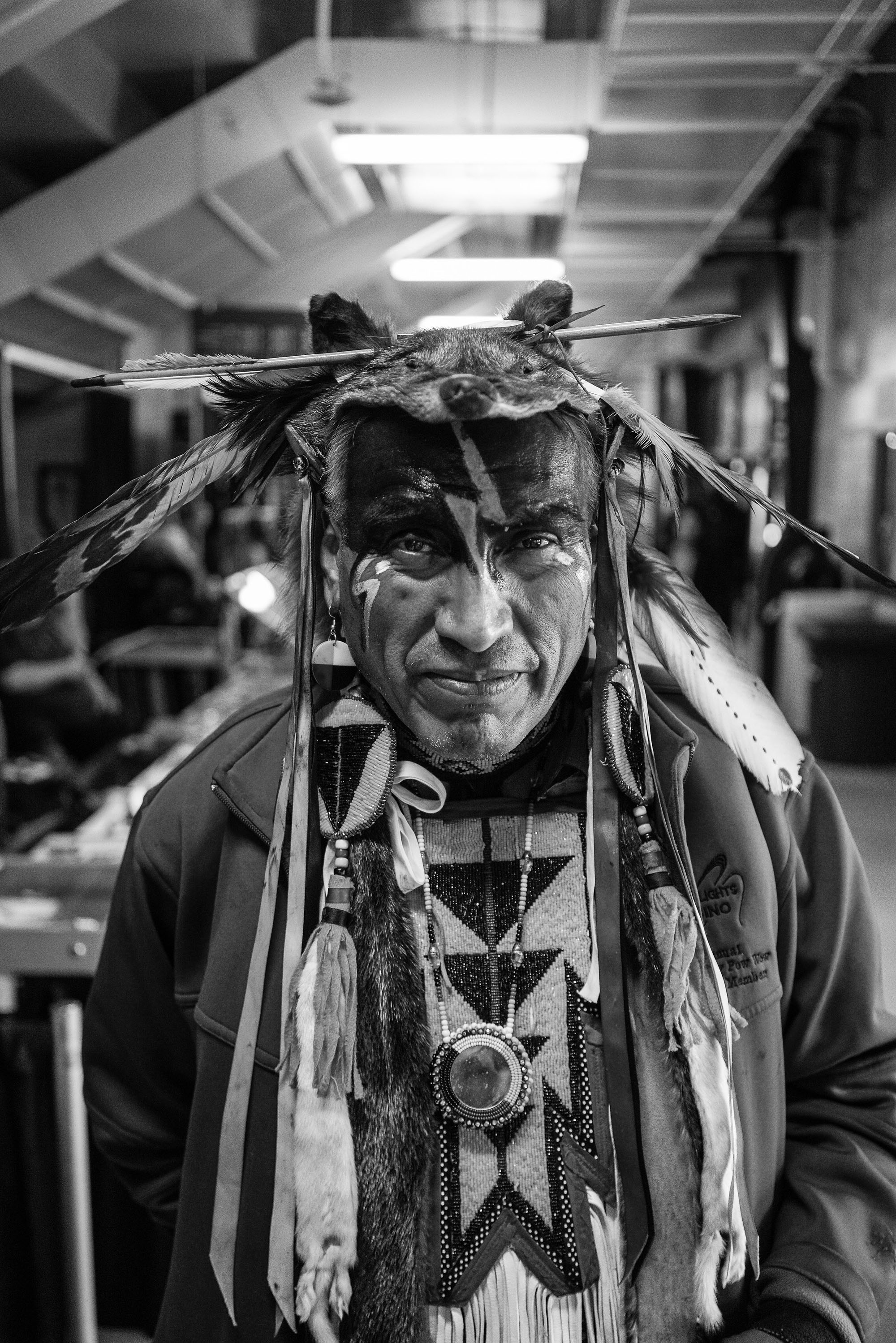 Black and white portrait of a man wearing traditional feathered headdress and ceremonial attire indoors