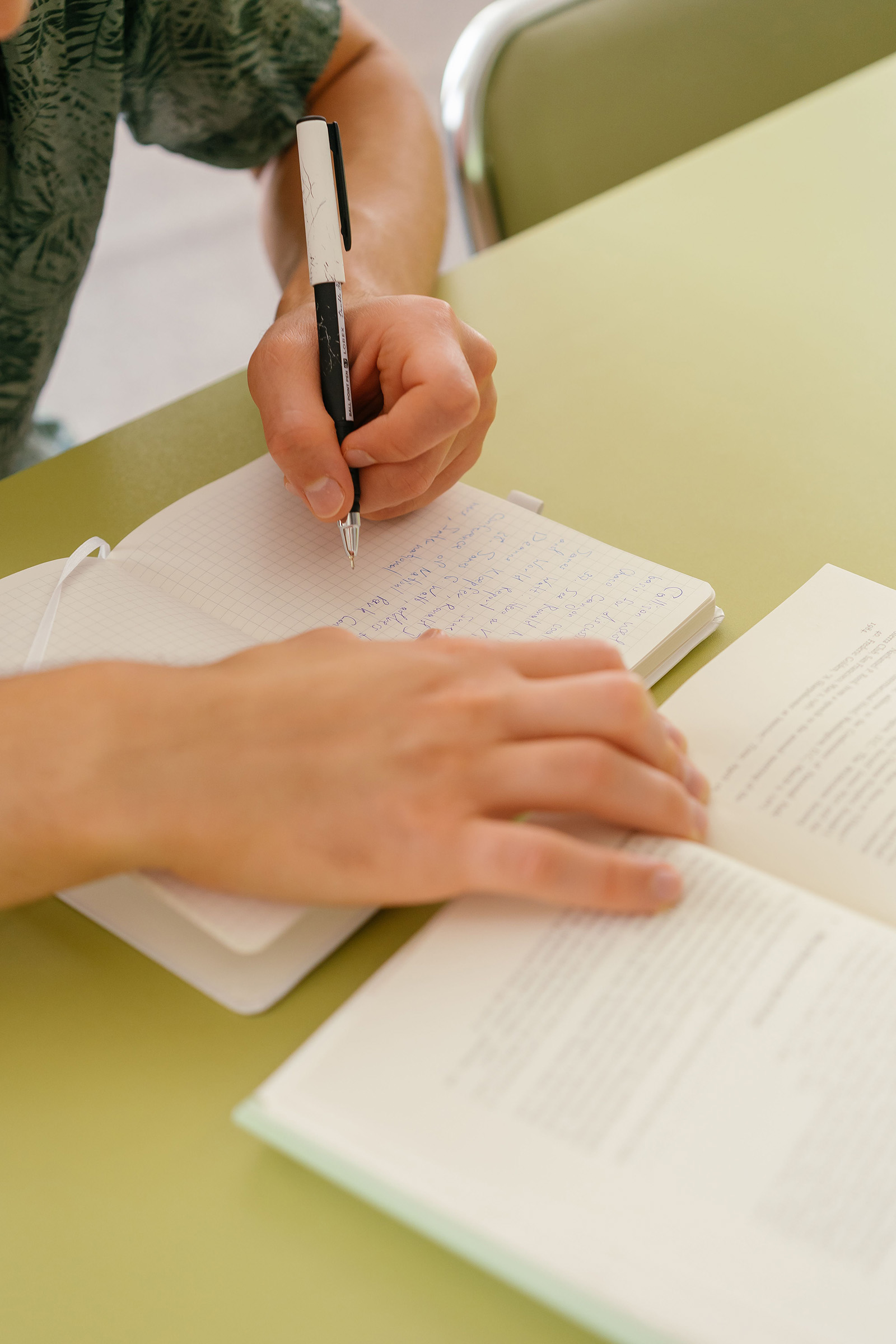 Close-up on student hands, left holding a pen and writing on notebook while right is holding pages of a book