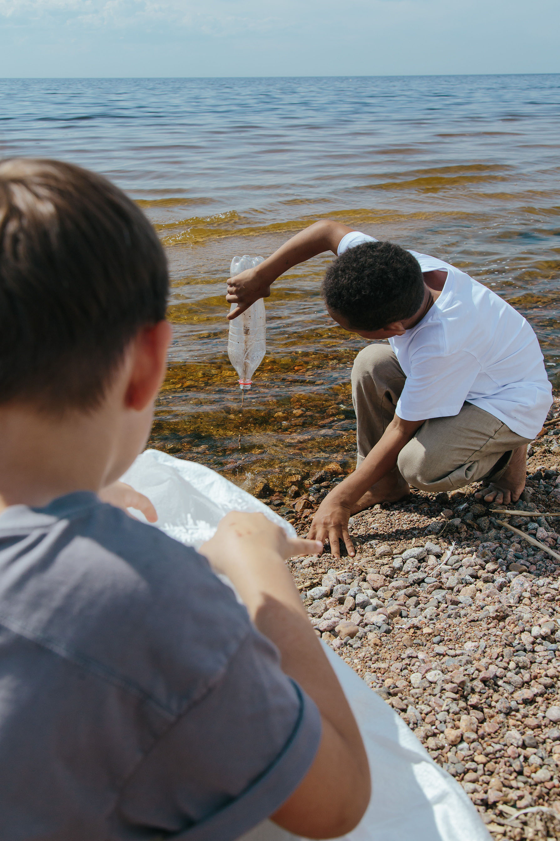 Two grade kids on the beach collecting trash, one is squatting close to water and holds a plastic bottle upside down